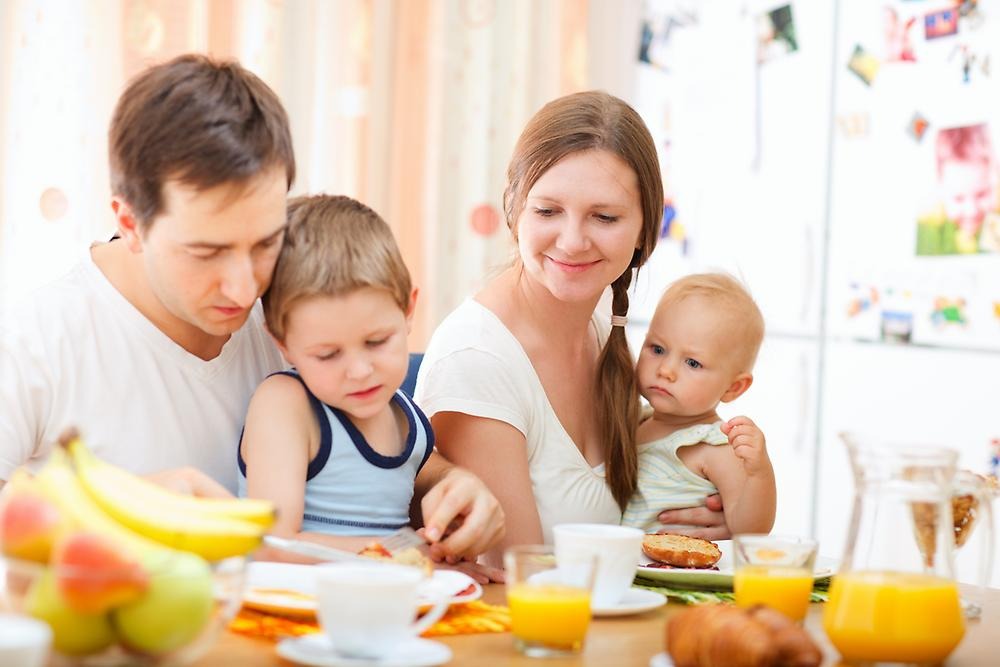 En familj sitter vid frukostbordet. Mamman och pappan har barnen i knät. På bordet finns frukt, smörgåsar och apelsinjuice. 