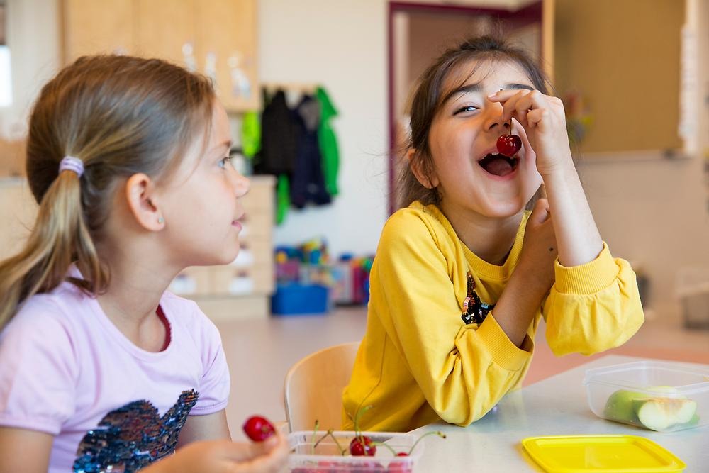 Två flickor sitter bredvid varandra i klassrummet. Den ena flickan skrattar och äter ett körsbär.